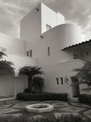 Black and white image of Von Romberg House, courtyard angle, white building, stone centre piece, with star design stone floor, palm tees, shrubs and hedges, archway with stone column, outside lantern light, ground and upper floor slim windows