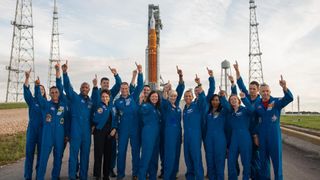 a bunch of astronauts in flight suits standing in front of a huge rocket and pointing at the sky