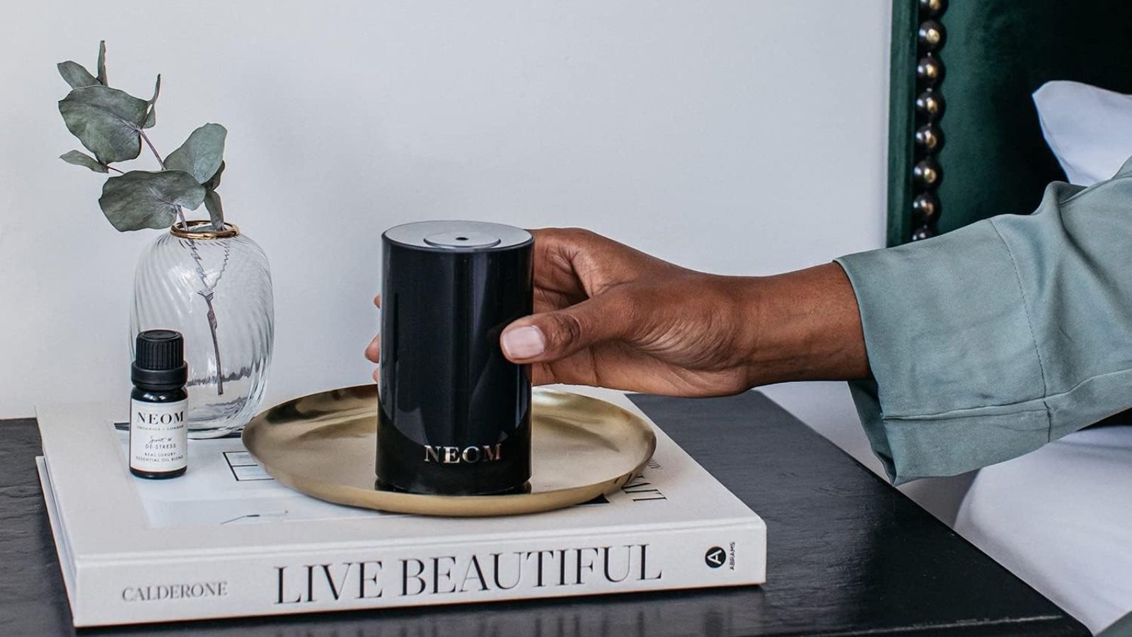 NEOM Wellbeing Pod in black on stack of books with hand holding it