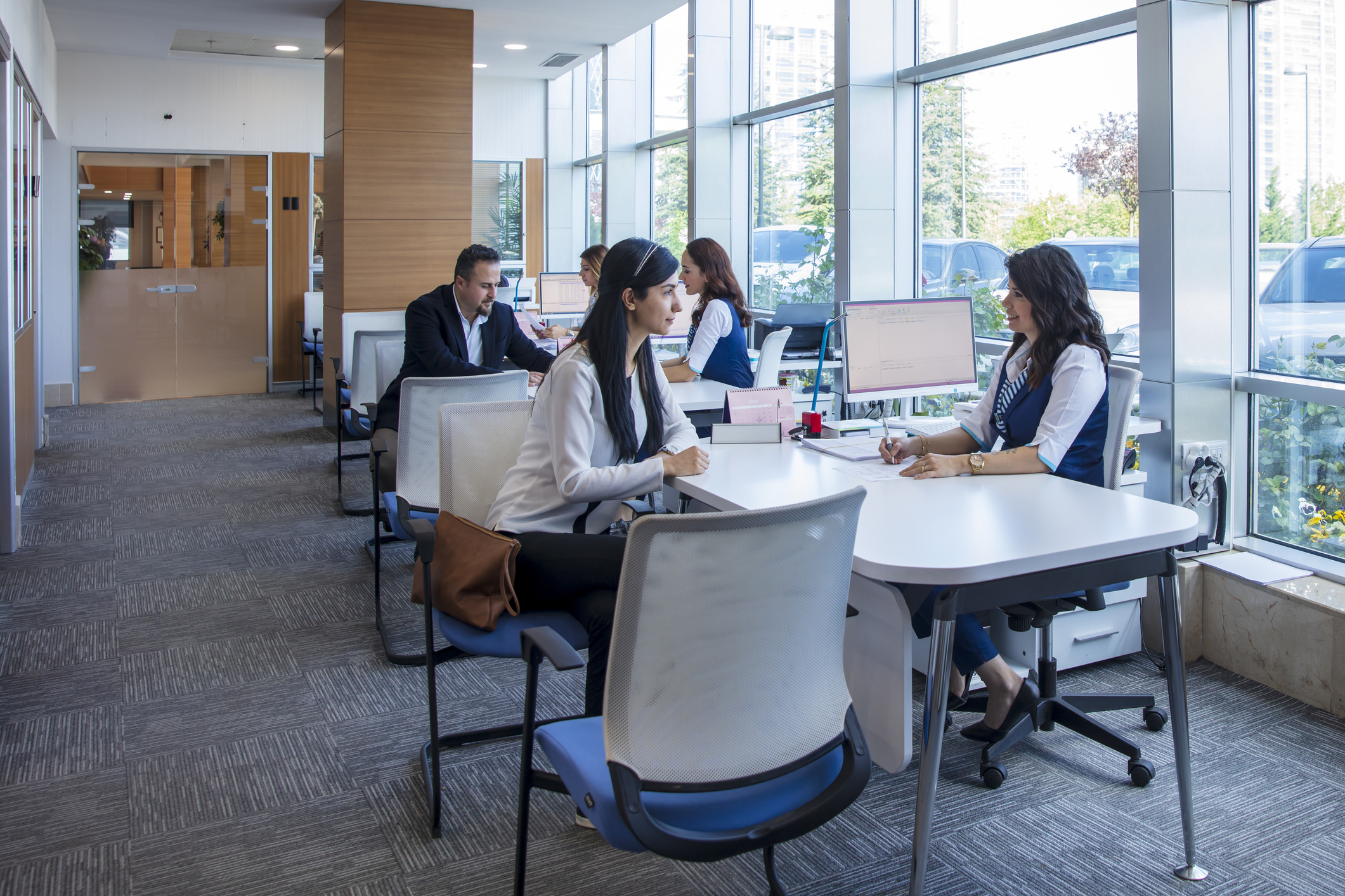Employees helping customers at a banking building with windows to the outside on a sunny day.