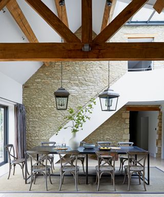 Large dining room with double height vaulted ceiling, dark wooden beams, stone wall, large dark wood dining table with nine dining chairs, two black metal and glass lantern style pendant lights, rug on the floor to zone the space