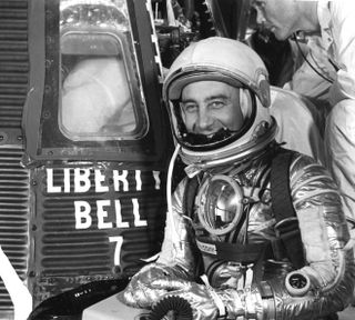 a man wearing a spacesuit smiles and sits next to a spacecraft with Liberty Bell 7 written on the side of it.