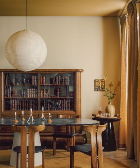 A modern dining room with a neutral wall, dark yellow ceiling, a large vintage wooden bookcase, a modern black dining table and wooden chairs, an ochre rug, and ochre curtains. 