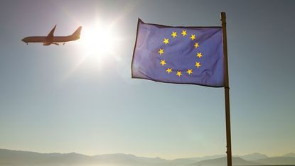 A European Union flag flies on a shore with mountains in the background and a plane flying overhead in the sunlight.