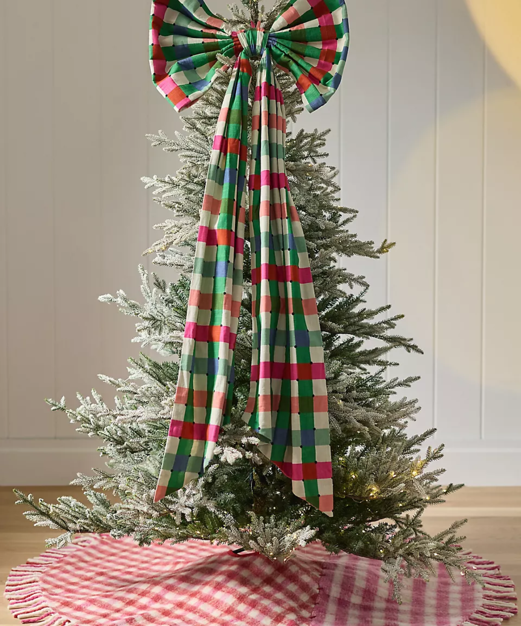 Anthropologie giant plaid bow tree topper crowning the top of a faux frosted Christmas tree, grounded by a wool-and-cotton-blend patchwork plaid skirt.