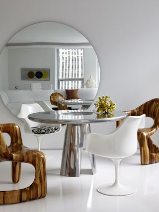 Image of an all-white dining room area. There is a chrome metallic, circular table with two white chairs and two wooden funky chairs around it. On the wall behind the table is a large, round mirror.