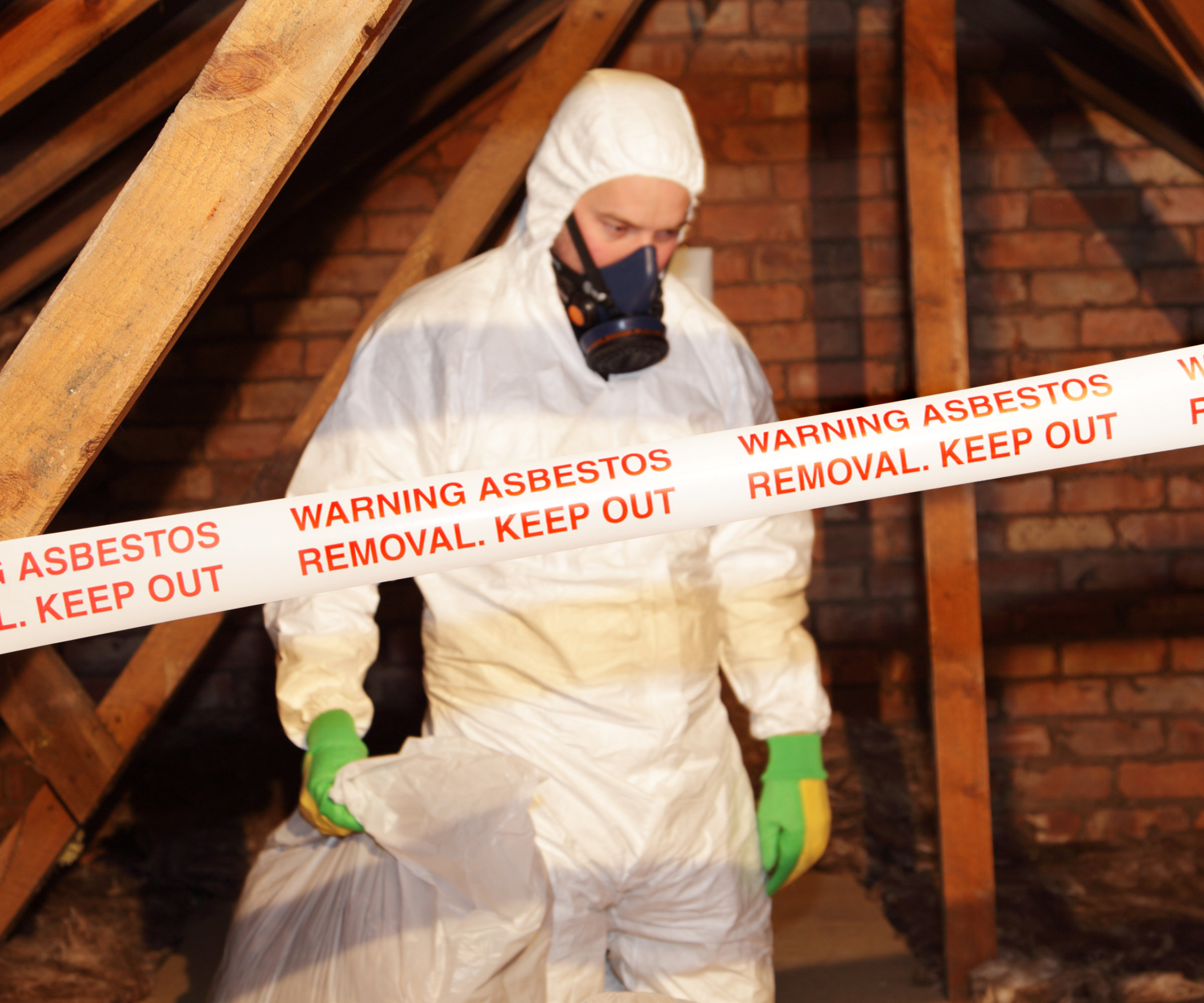 worker wearing protective white suit, mask and gloves removing asbestos from loft