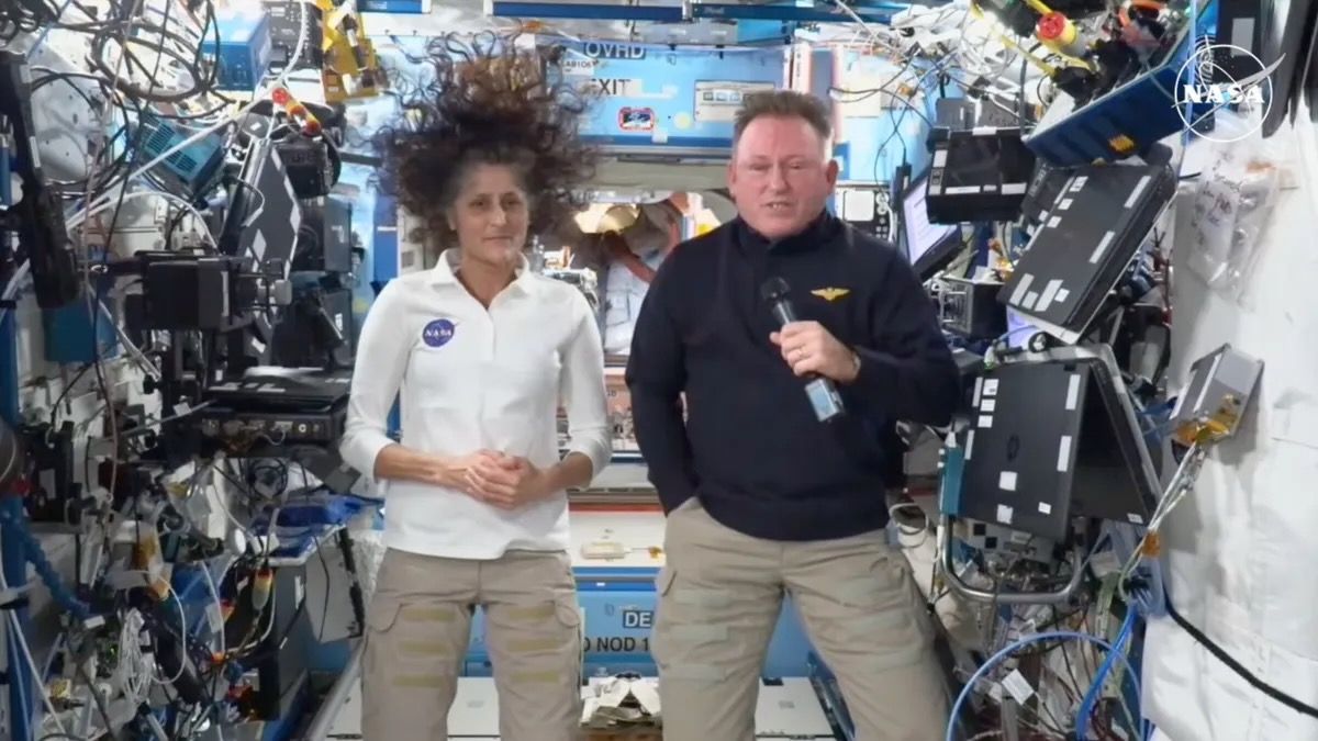 a man and a woman, both wearing khaki slacks, speak while aboard the international space station, with lots of wiring and computer equipment around them