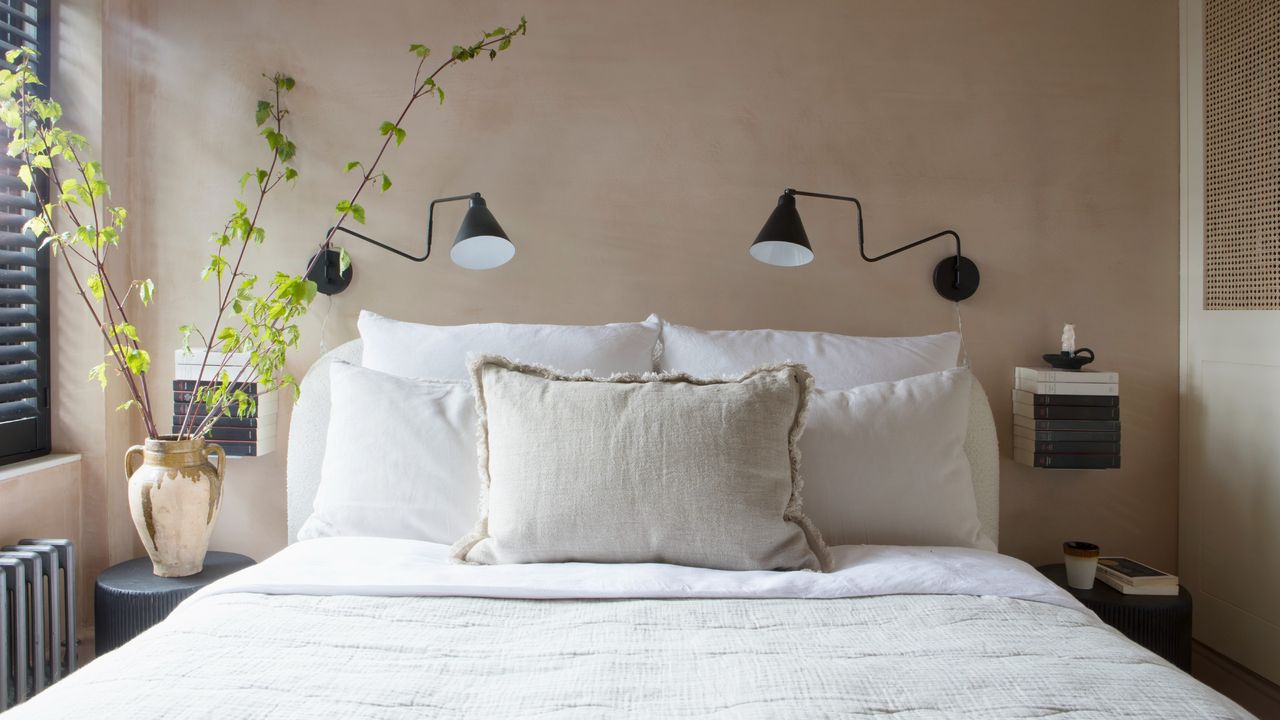 A bedroom painted in a peach shade with a limewash finish with black wall lights on the wall and a vase of leafy branches on the bedside table