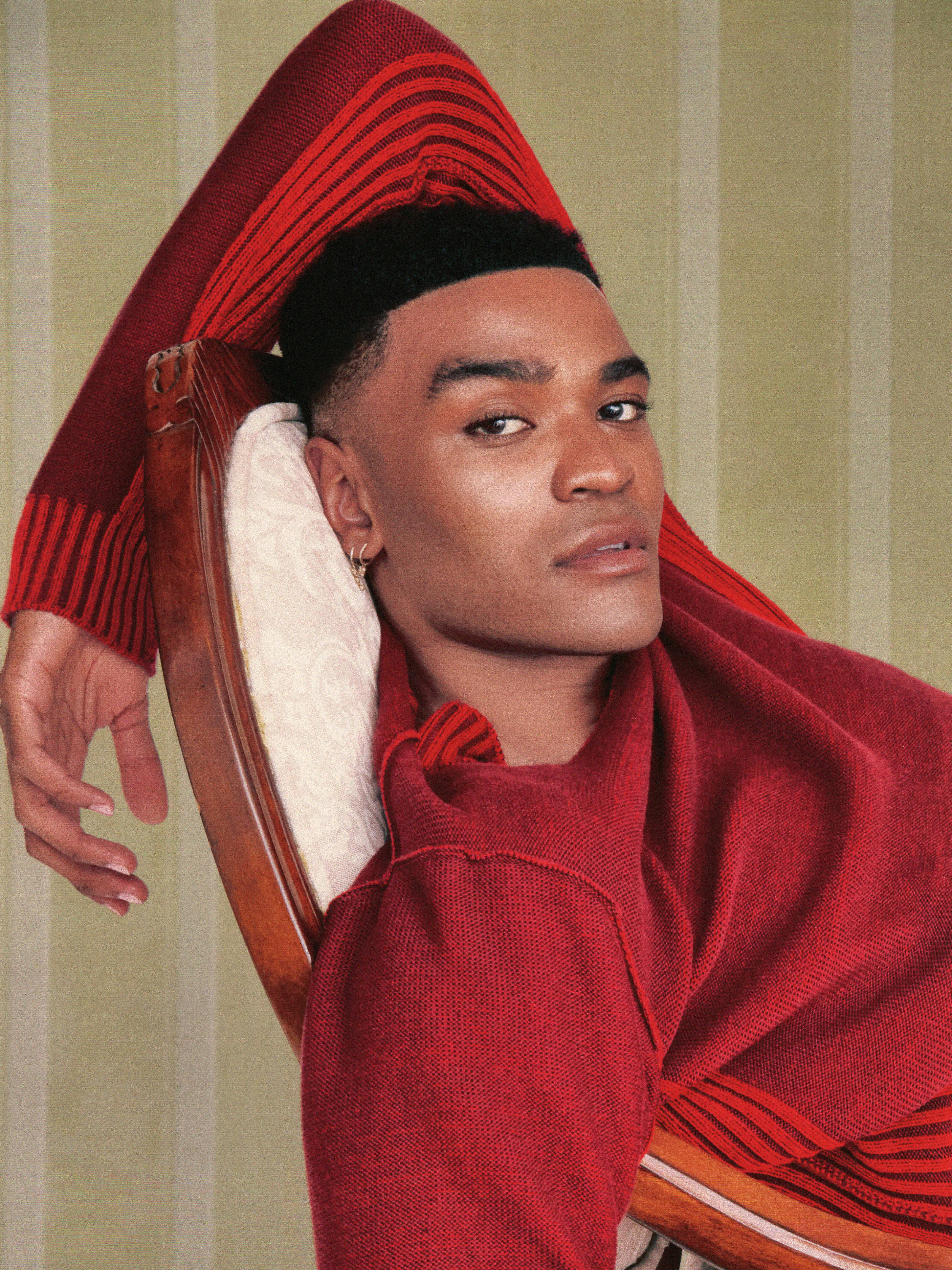 Owen Thiele poses leaning back in a vintage looking chair wearing a red sweater against a green striped backdrop