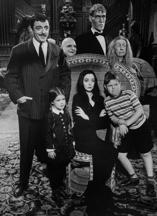 Carolyn Jones (C, sitting) and John Astin (L), with other cast members, sitting around a chair in a spooky house during scene from program The Addams Family.