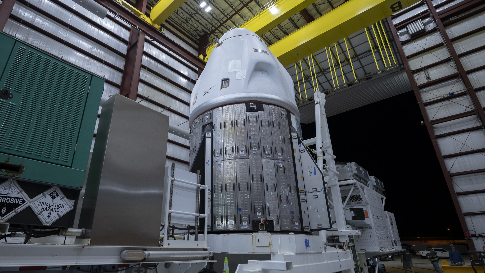 a conical white space capsule inside a hangar, with the night sky visible through a large open door behind it