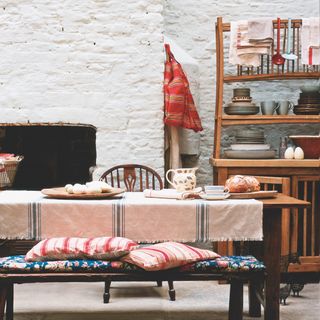 rustic kitchen with dresser full of tableware, large table with linen tablecloth, bench with floral and stripe cushions, painted brick wall, enamelware, jug, rustic platter