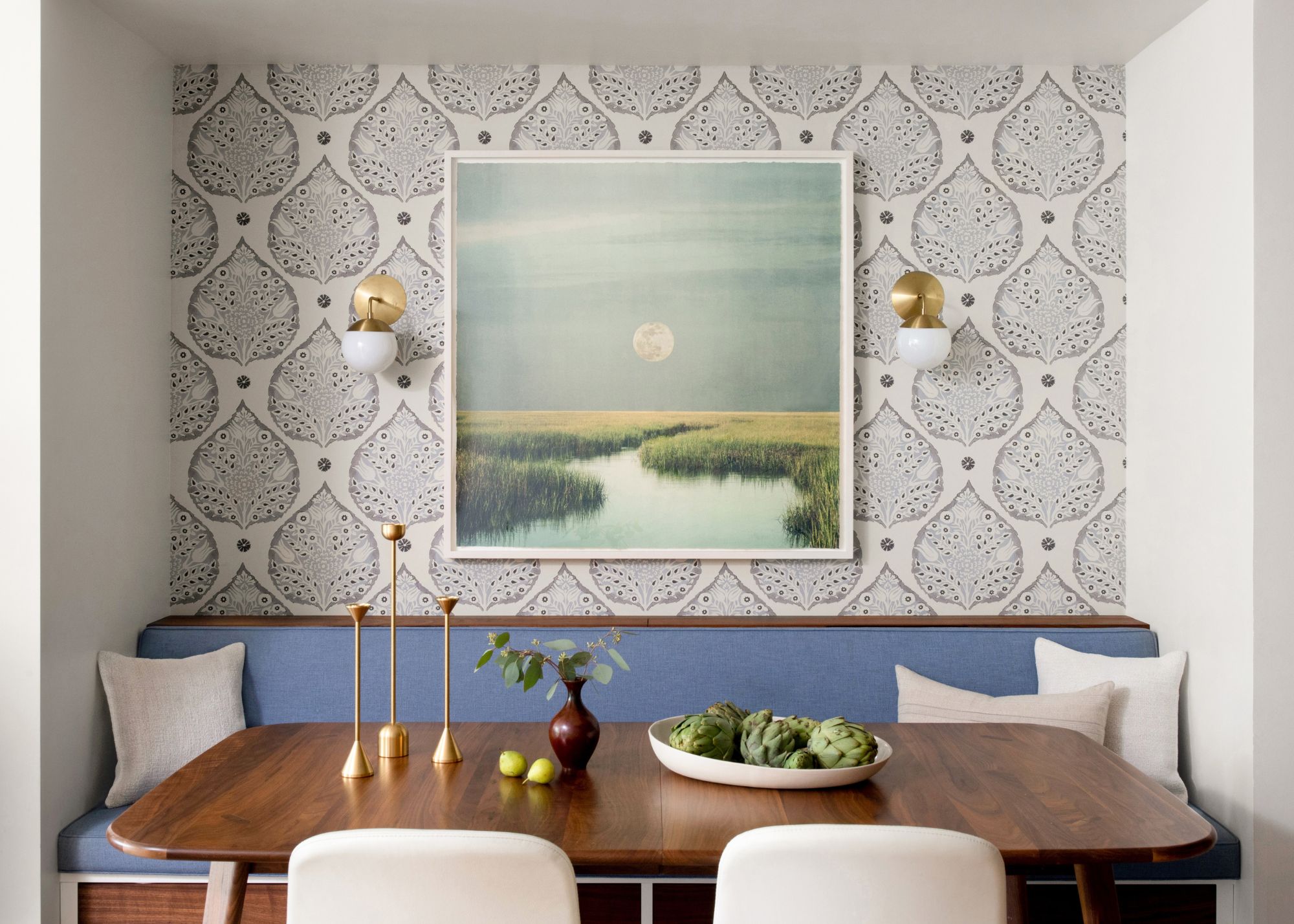 An apartment dining nook with neutral patterned wallpaper, powder blue banquette seating, a dark wood table, and light neutral cushions. A landscape artwork on the walls with two brass sconces.