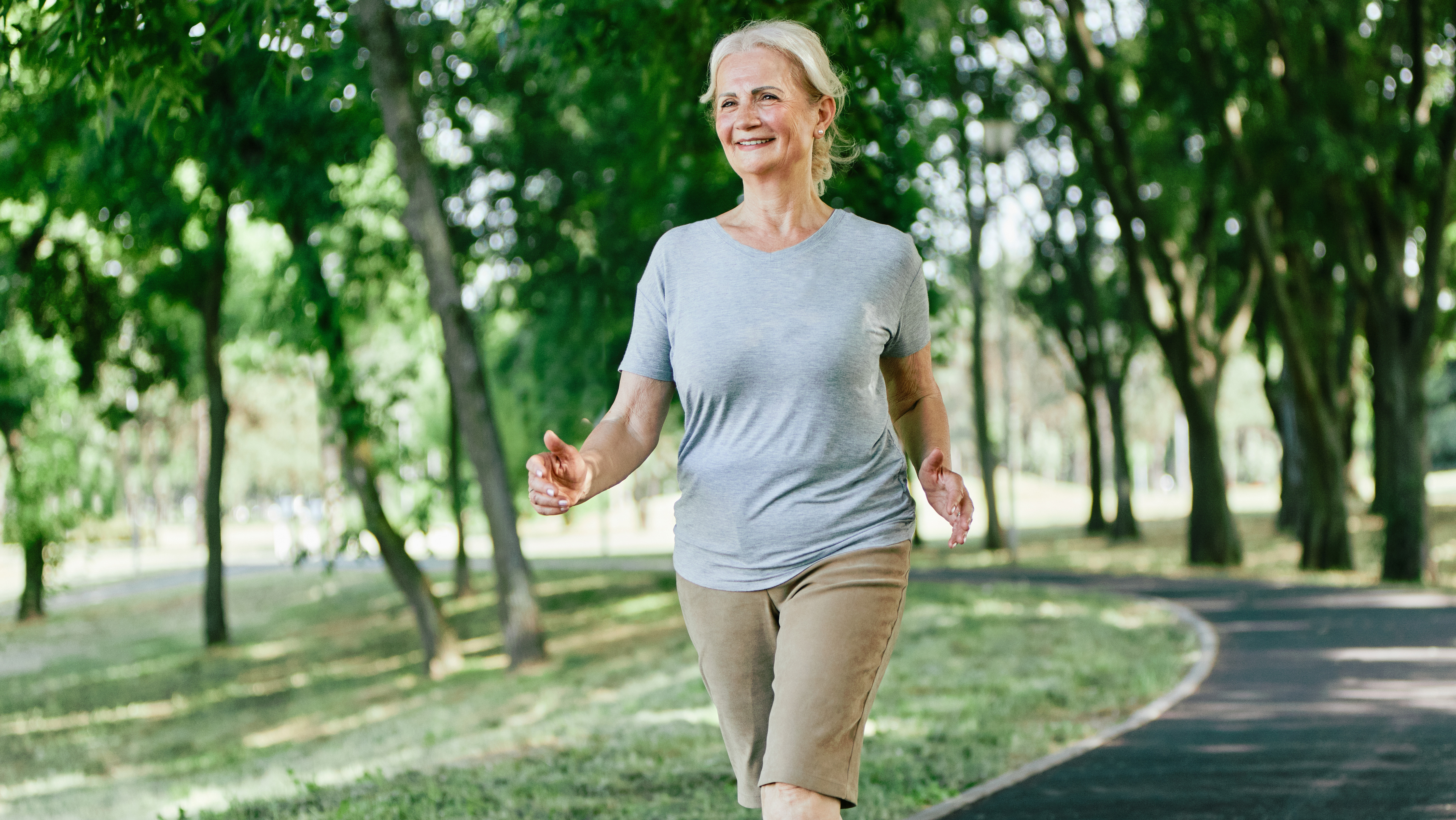 A woman power walks in a sunny park with trees.