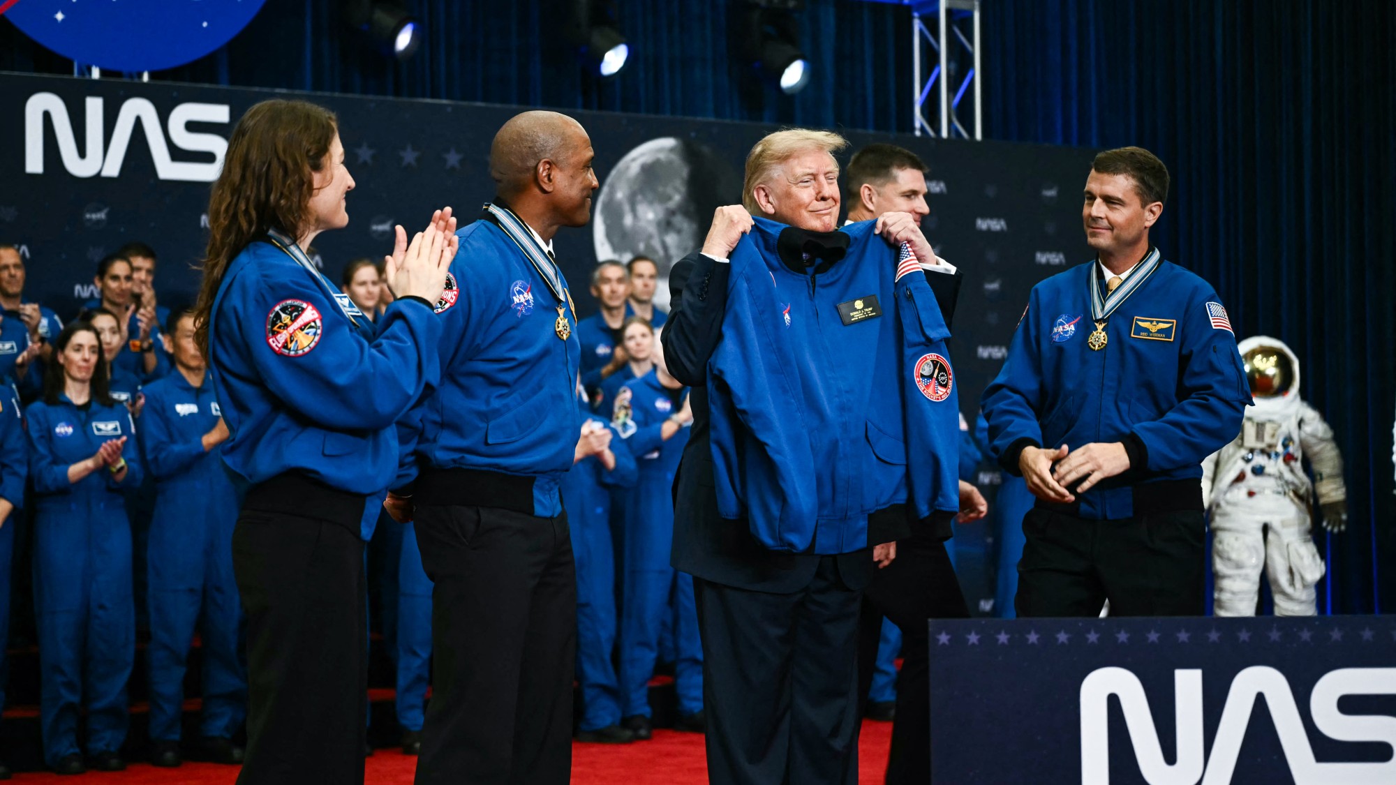 a man in a blue suit gives medals to four people in blue flight suits on a stage with a large sign that reads "NASA"