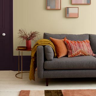 skirting board colour ideas, living room with pale green walls, artwork, white painted floor boards, coral cushions, turmeric throw, metallic side table, rug, deep violet skirtings and door, grey sofa