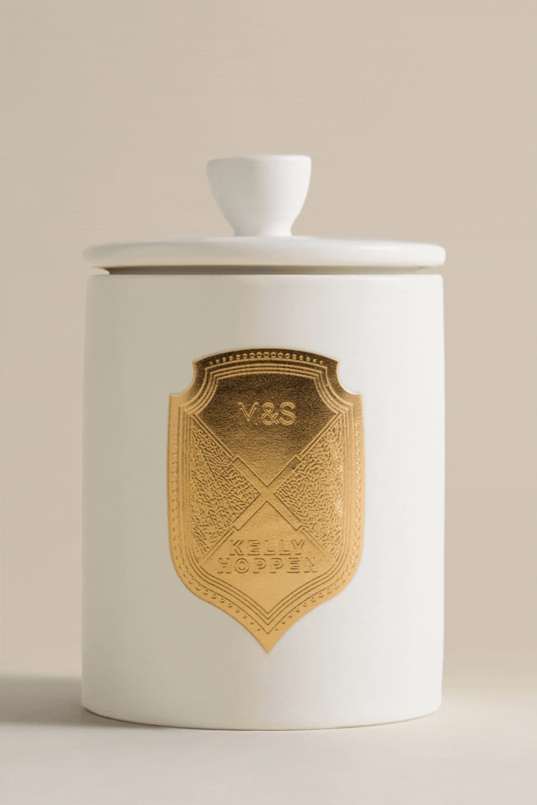 A GIF of a scented candle in a white ceramic vessel with a matching lid and a gold crest in the middle