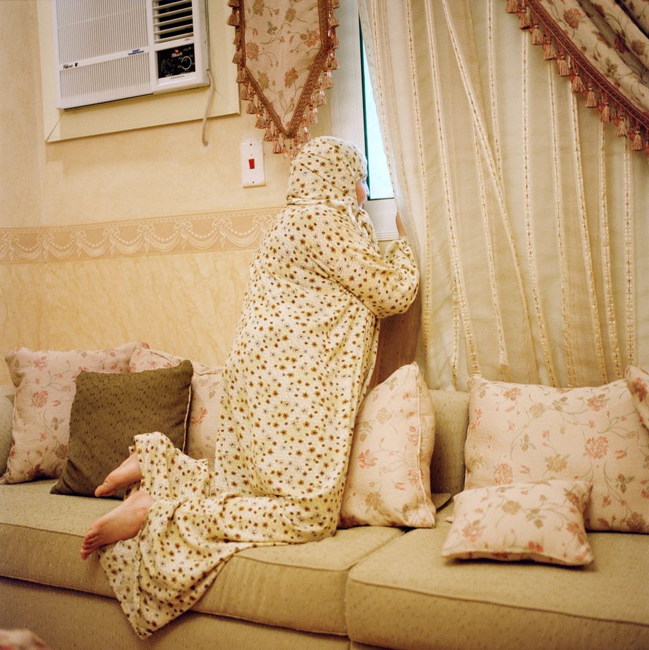 A woman wearing a prayer dress kneeling on a sofa peeking out a window through the curtains, with her dress and furnishings all in shades of brown and cream