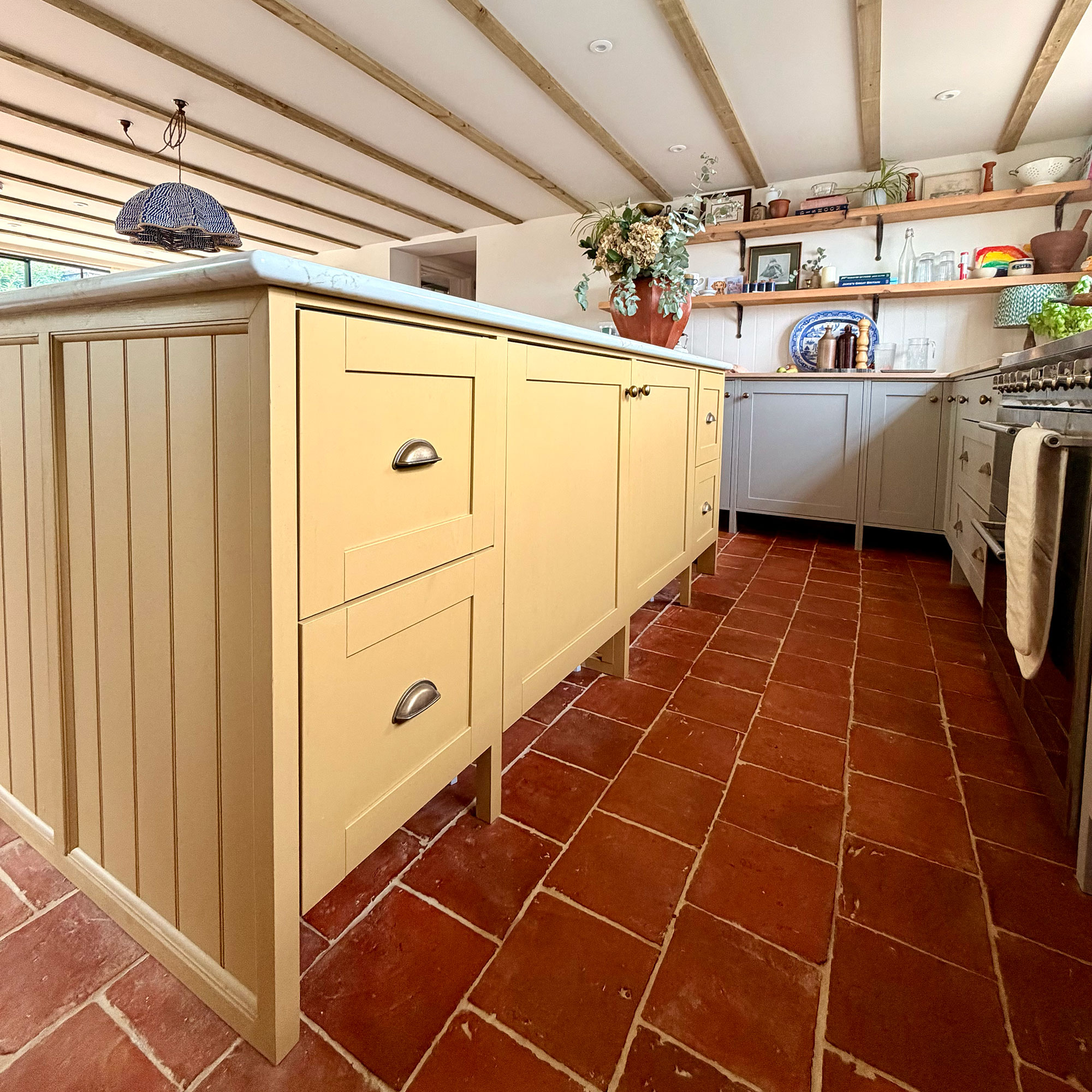 Kitchen with tiled terracotta flooring, wooden beams in ceiling and butter yellow island