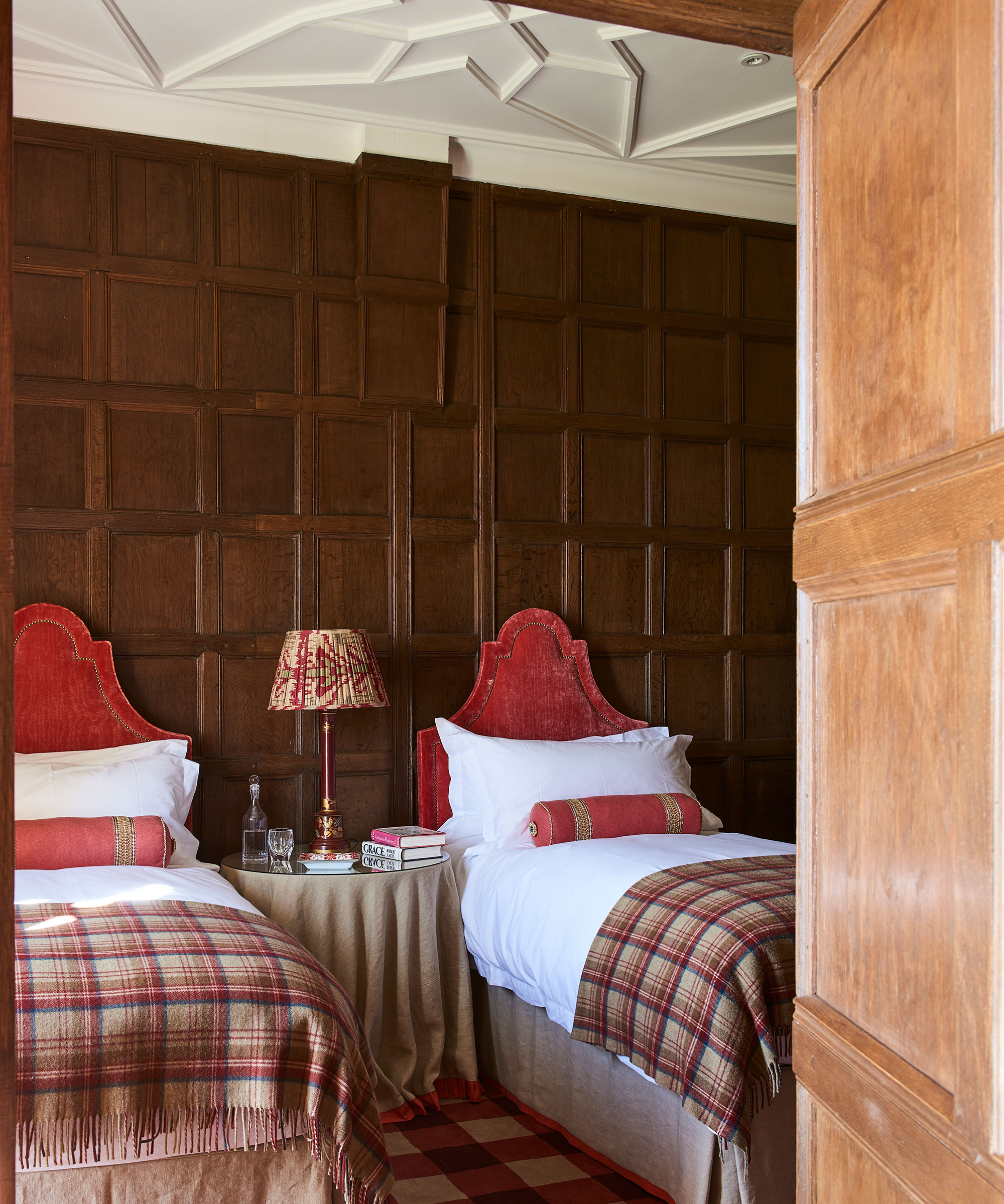 Small bedroom with twin beds with red headboards and tartan throws in a wood-panelled room.