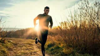 A man wearing black lycra running through field with rising sun shining behind him