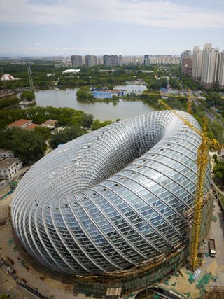 View from above of the irregular, ring shaped Phoenix TV building during the day. There are two yellow cranes next to the building and there are other buildings, greenery and a body of water nearby