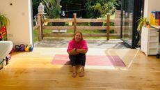 Fitness writer and journalist Maddy Biddulph sits on a wooden floor at home and smiles at the camera. Behind her we see a backyard and large, open glass doors.