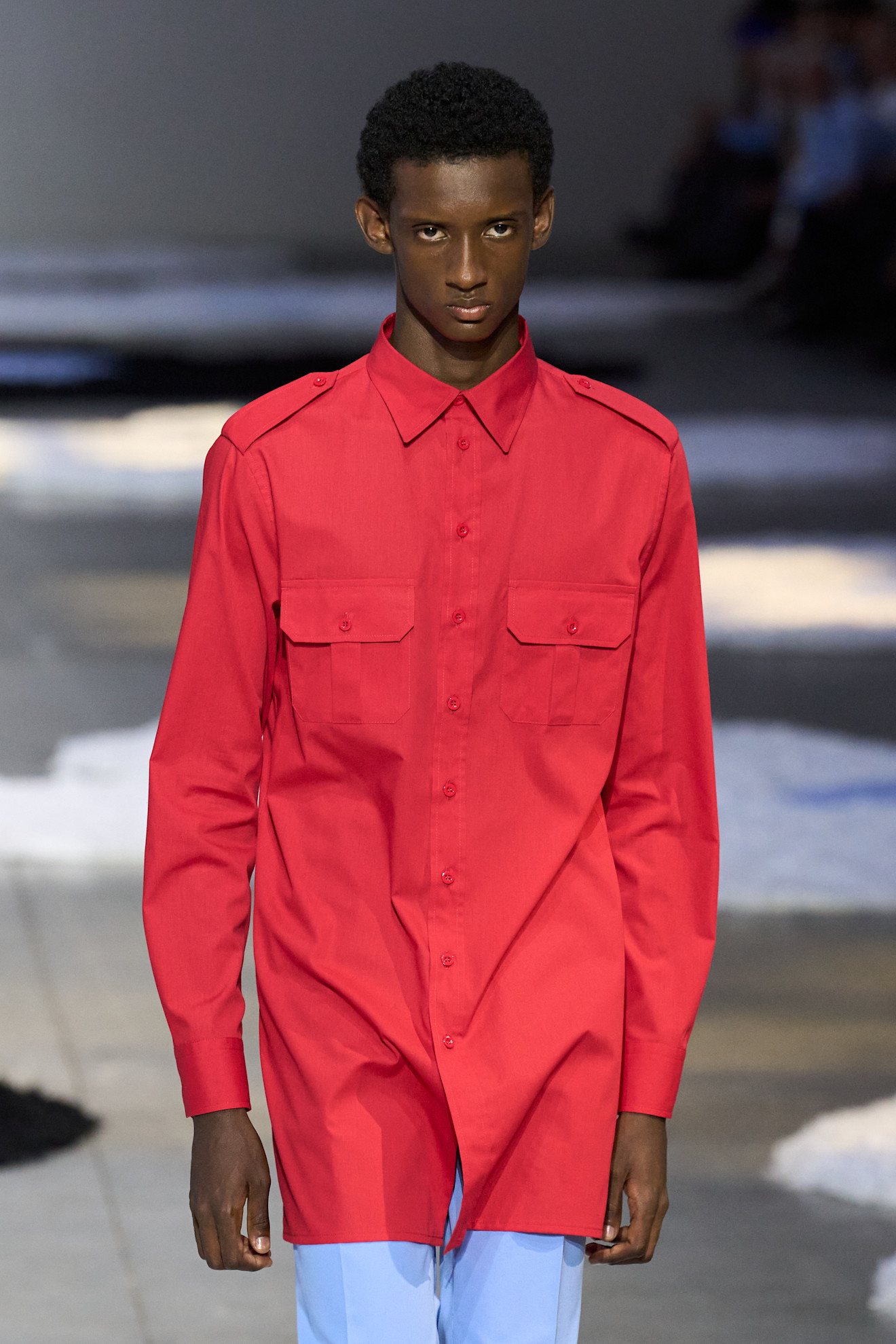 A man wears a red button-down shirt with blue trousers.