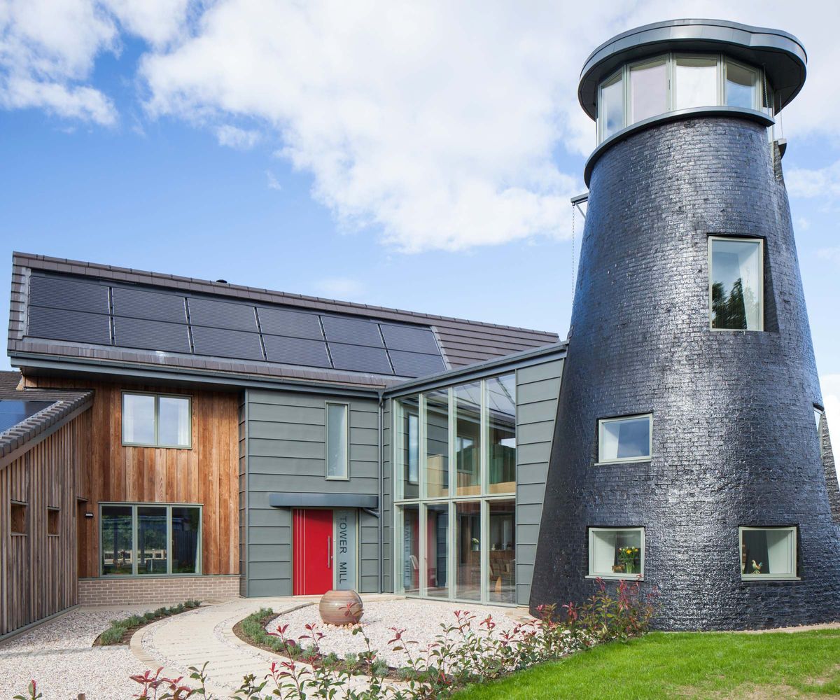 A self build home connected to a circular water tower-like structure to one side. The large house features timber cladding and solar panels on the roof. The large windows are from VELFAC.