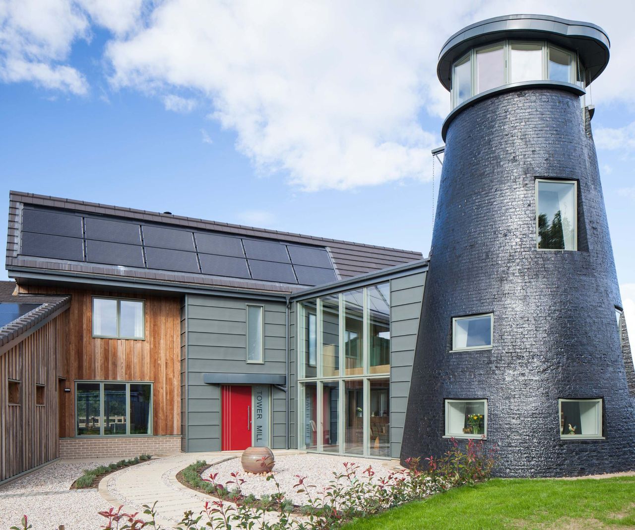 A self build home connected to a circular water tower-like structure to one side. The large house features timber cladding and solar panels on the roof. The large windows are from VELFAC.