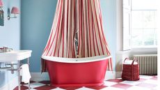 Bright cornflower blue and red circus theme bathroom with Victorian sink style, checkered red and white floor, retro radiator, shutters, red freestanding bath tub with red and white shower curtain