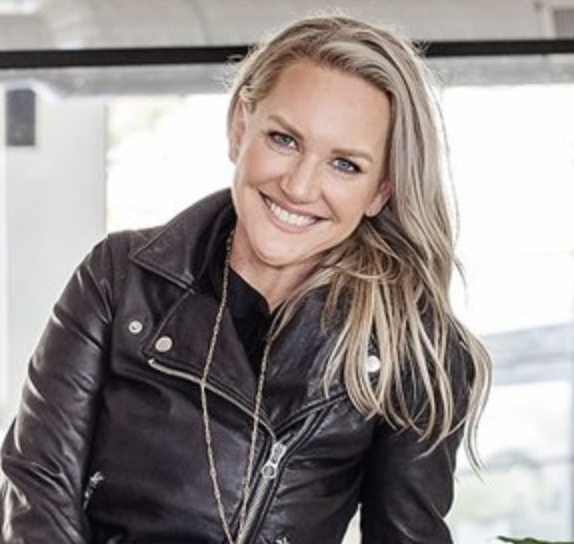 A headshot of a woman with blonde hair wearing a leather jacket and smiling at the camera