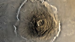 A close up of Olympus Mons on the surface of Mars, its structure a pile of brown lava in a large circular mount on the surface of the planet