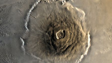 A close up of Olympus Mons on the surface of Mars, its structure a pile of brown lava in a large circular mount on the surface of the planet