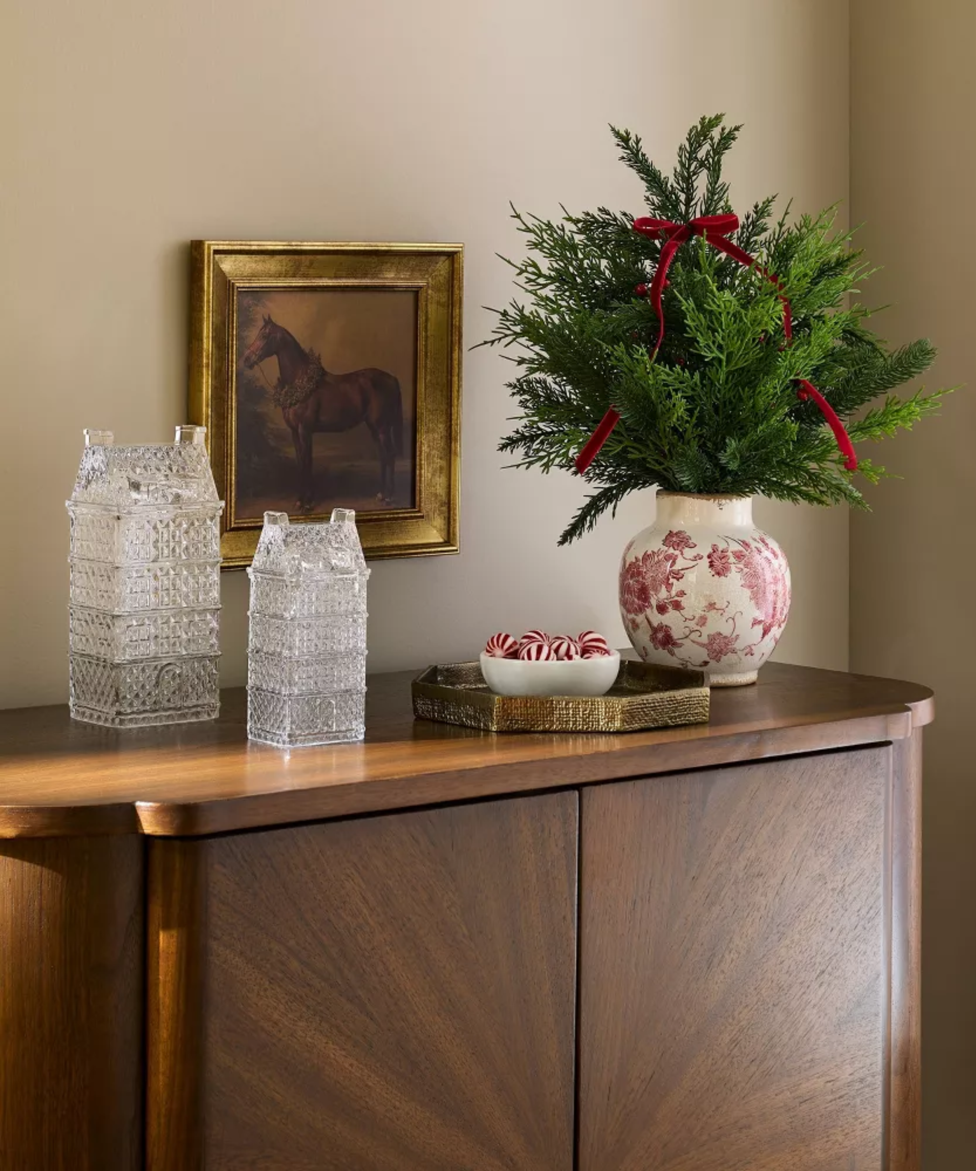 Holiday bar cabinet vignette featuring etched glass holiday house figurines alongside a golden decorative tray, potted evergreen plant, and equestrian wall art.