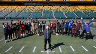 The Amazing Race Canada presenter standing in front of the season 11 cast in an American Football stadium.