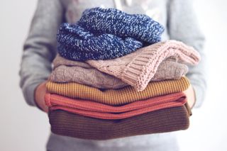 how to wash clothes by hand. The mid-section of a person in a gray sweater holding a pile of autumnal colored clothing