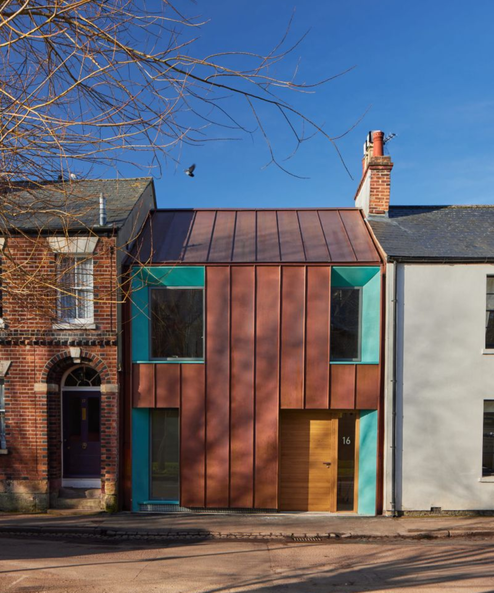 terraced property with copper clad frontage and sloped, green metal window and door reveals and timber door nestled between a brick home on one side, white rendered property on the other