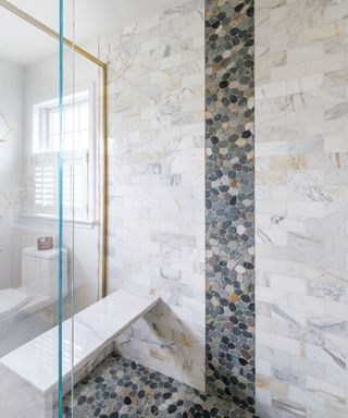 A white bathroom shower with marble subway tiles and a strip of gray stone tiles in the middle cascading onto the floor, and a marble shelf in the bathroom