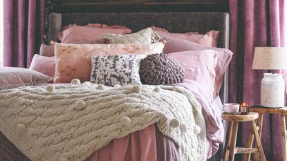 A cosy bed in a pink colour scheme with many pillows and scatter cushions on top and a knitted cream throw, all framed with pink velvet curtains on either side