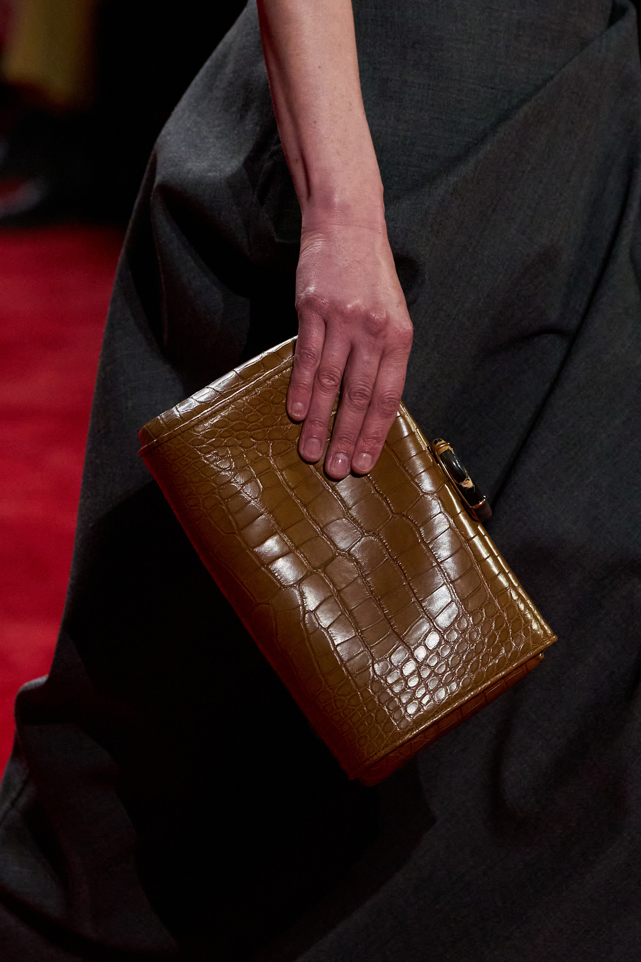 A close-up photo of a model walking in the F/W 26 Bottega Veneta show carrying a brown croc clutch.