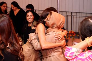 Meghan Markle hugging a woman wearing a tan head scarf as another woman watches on smiling