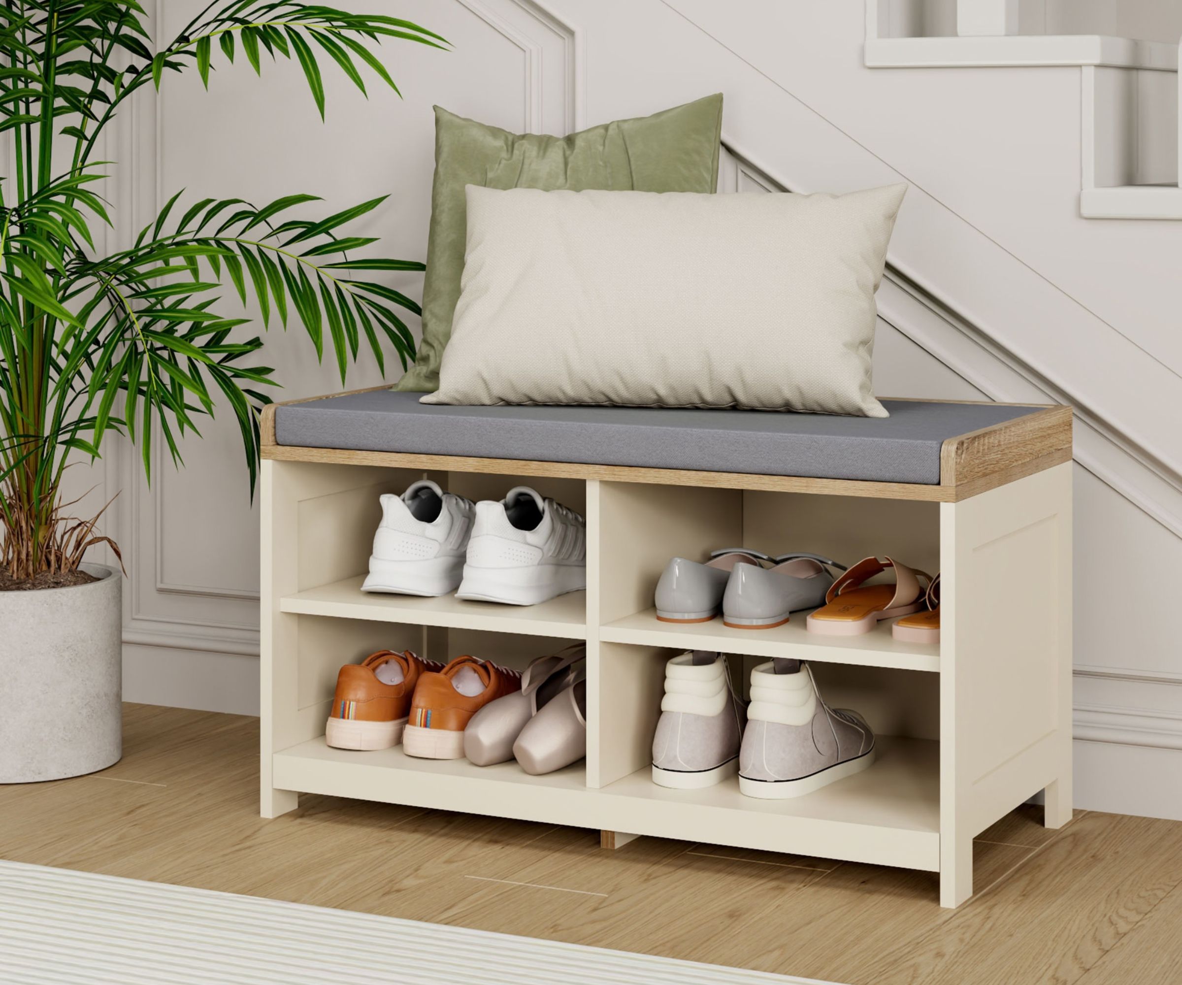 A cream and oak shoe storage bench with four open cubby compartments holding trainers, sandals and casual shoes, topped with a grey upholstered cushion and two scatter cushions, positioned at the foot of a staircase beside a potted palm.