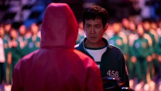 Promotional image of &#039;Squid Game&#039; season 2 which shows Seong Gi-hun (Lee Jung-jae) standing up to a guard in front of a large group of contestants.