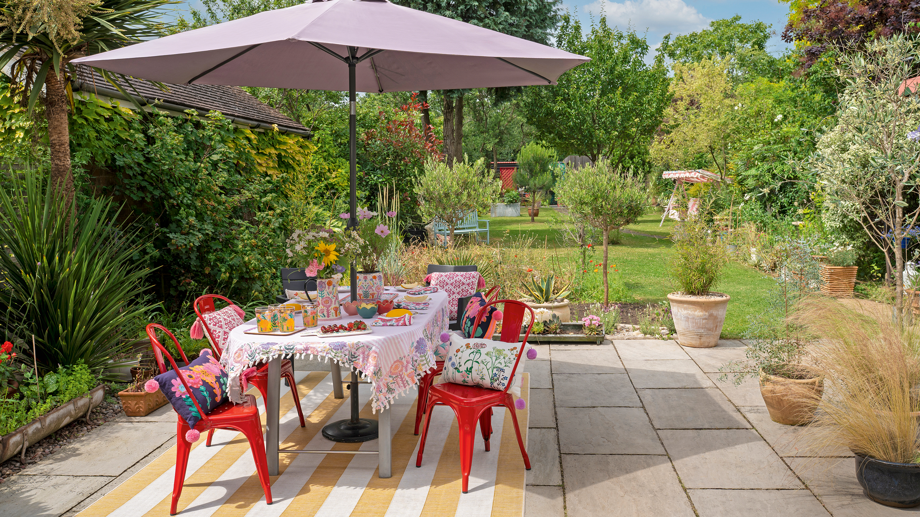 garden with a dining table and red metal chairs and parasol with a yellow and white striped rug