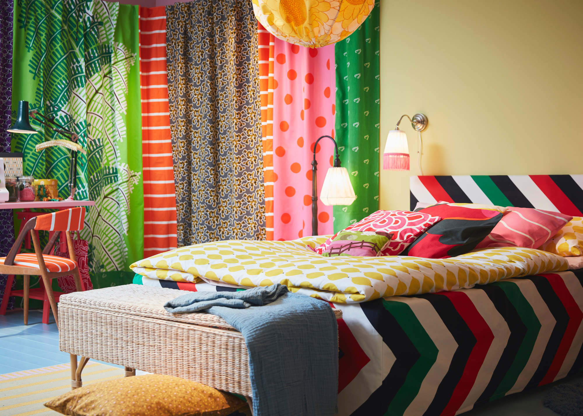A bright yellow bedroom with several colorful textiles hung from the ceiling, a floral pendant lantern above a bed dressed in vibrant black, red, white and green stripes, a yellow duvet and colorful pillows. A woven bedstand sits at the foot of the bed with a blue throw draped over and a deep yellow pillow on the ground beside it. An orange and white striped chair sits in the corner near a desk