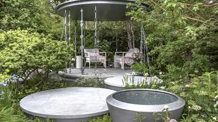 A show garden at the Chelsea Flower Show with a pergola, shrubs, and trees to provide shade