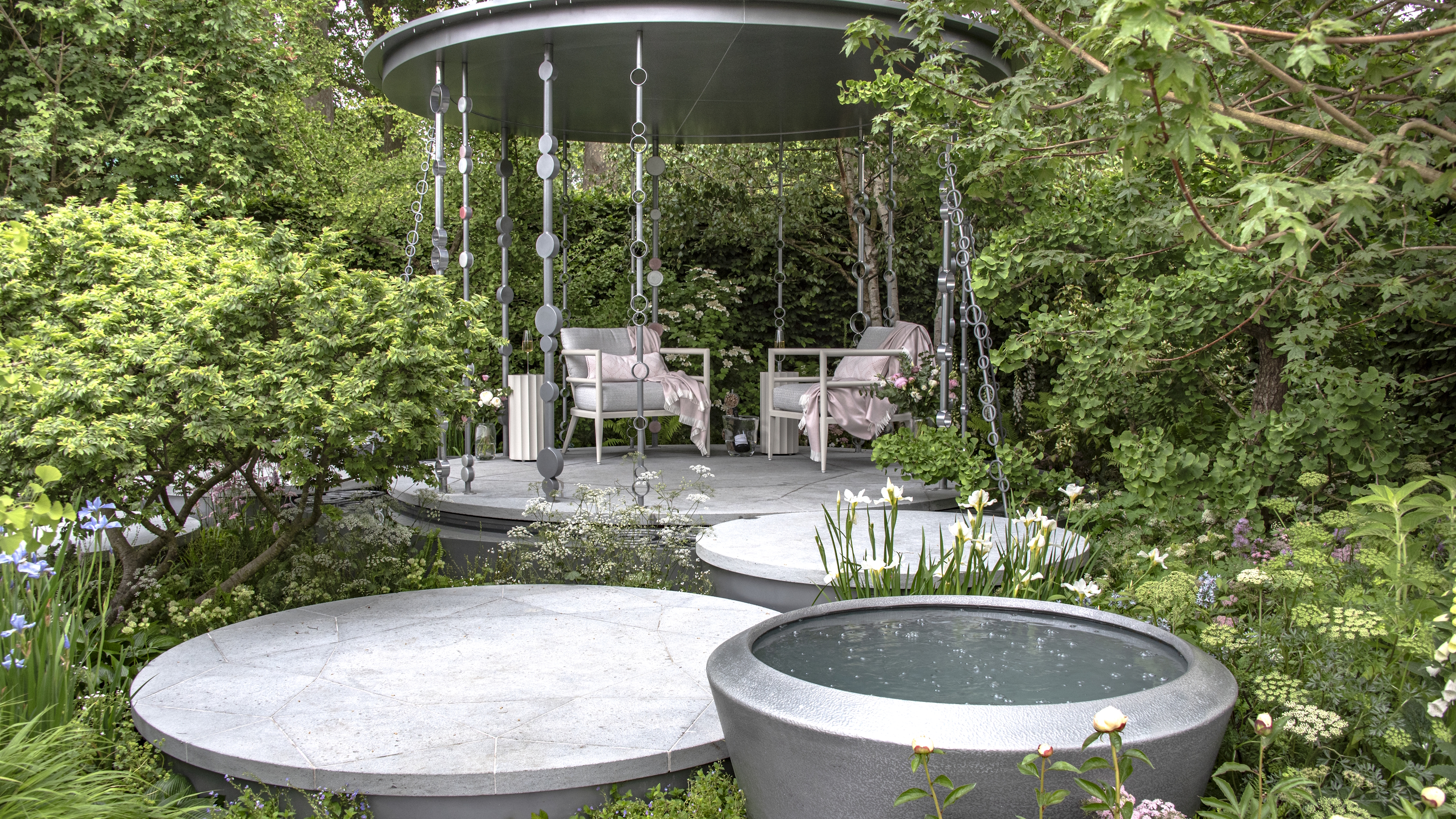 A show garden at the Chelsea Flower Show with a pergola, shrubs, and trees to provide shade