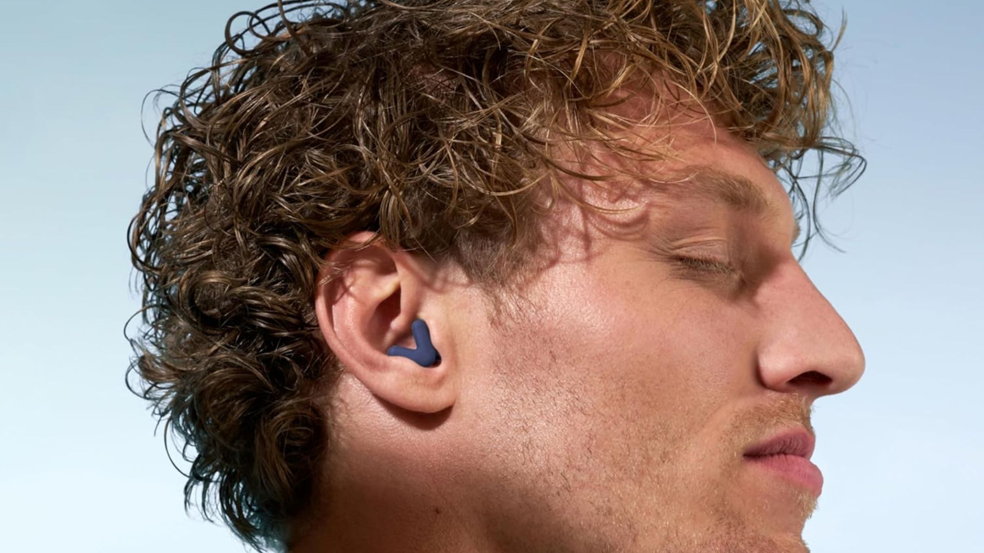 A man with blonde curly hair wearing a blue pair of Alpine Silence Earplugs